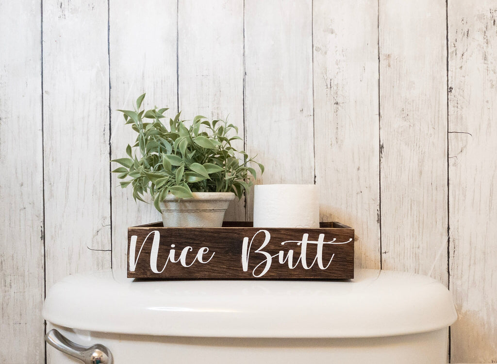 Nice Butt dark stained farmhouse toilet paper box on toilet tank with plant and toilet paper roll angled view