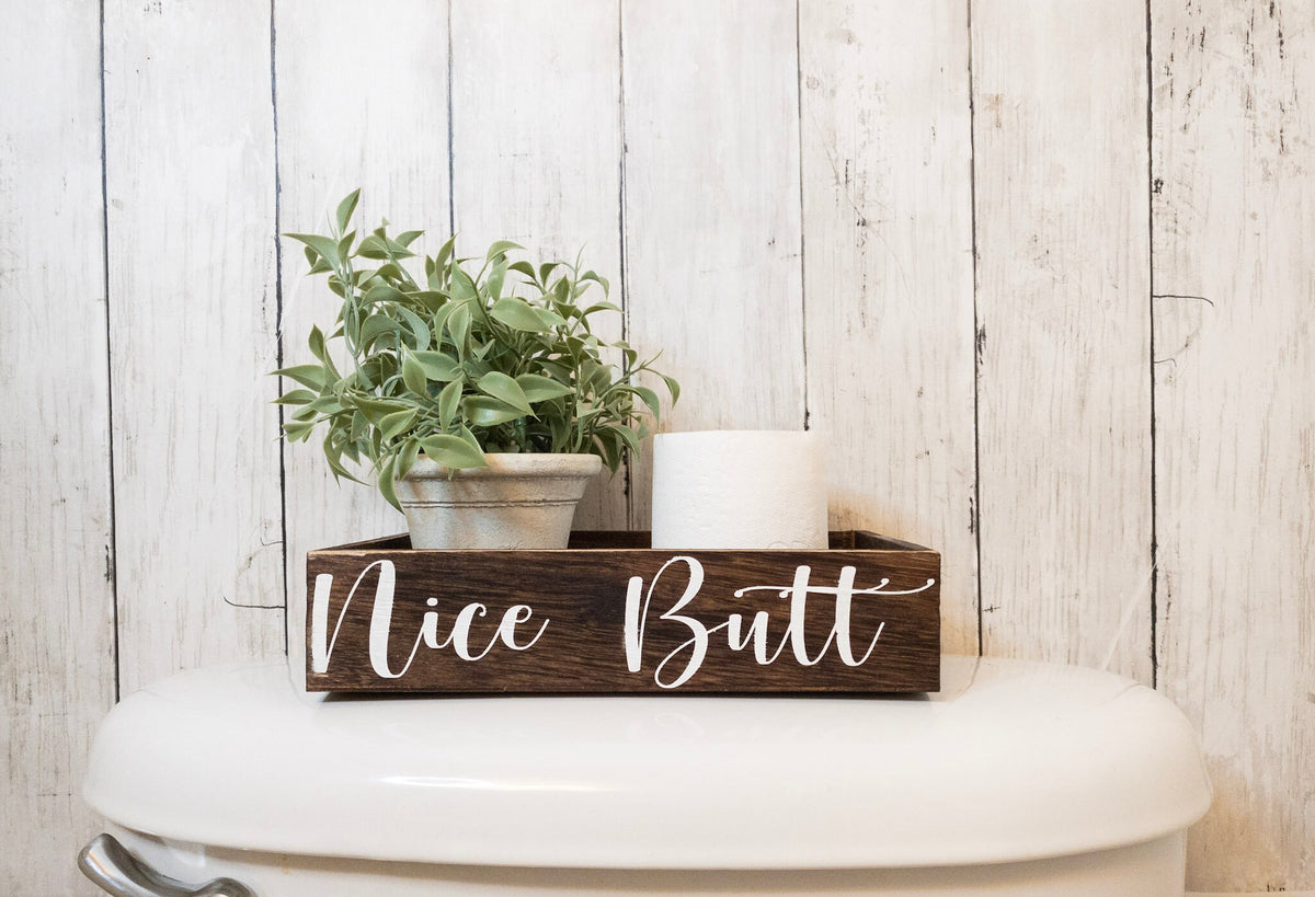 Nice Butt dark stained farmhouse toilet paper box on toilet tank with plant and toilet paper roll front view