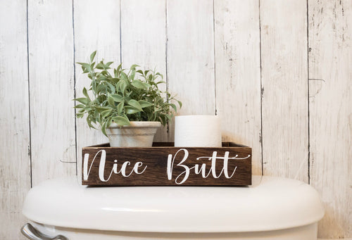 Nice Butt dark stained farmhouse toilet paper box on toilet tank with plant and toilet paper roll front view