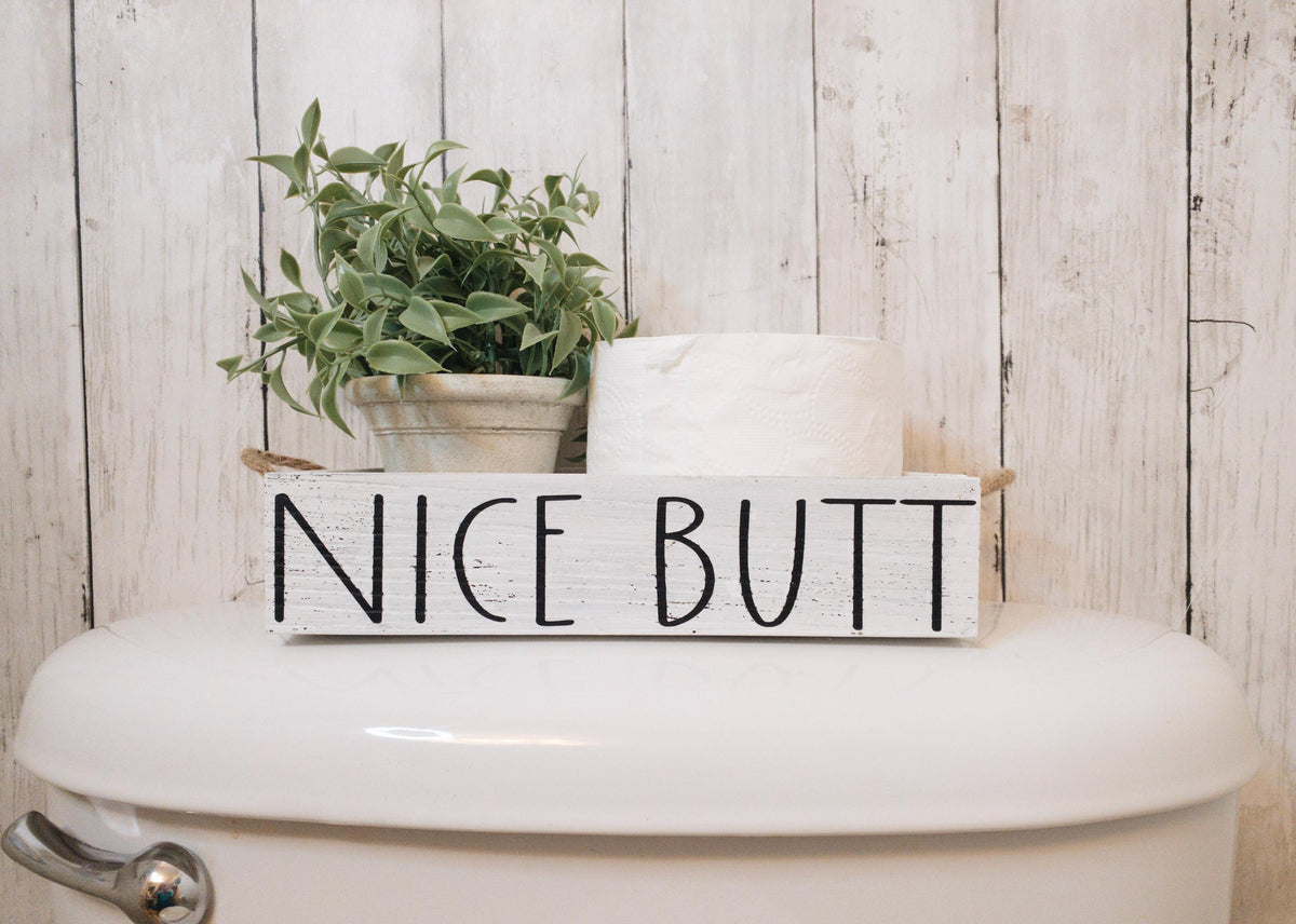 Nice Butt white farmhouse toilet paper box on toilet tank with plant and toilet paper roll close up