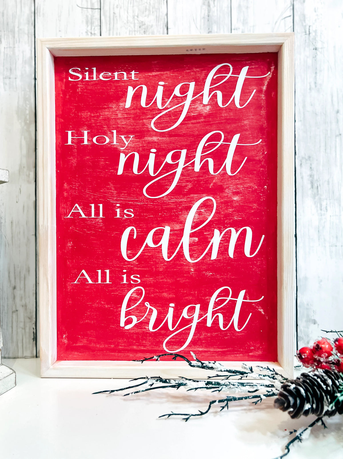 Red Silent Night Holy Night All is Calm All is Bright framed wood sign with whitewashed birch frame leaning against white shiplap wall with white lantern and red berry pine cone spray side angle