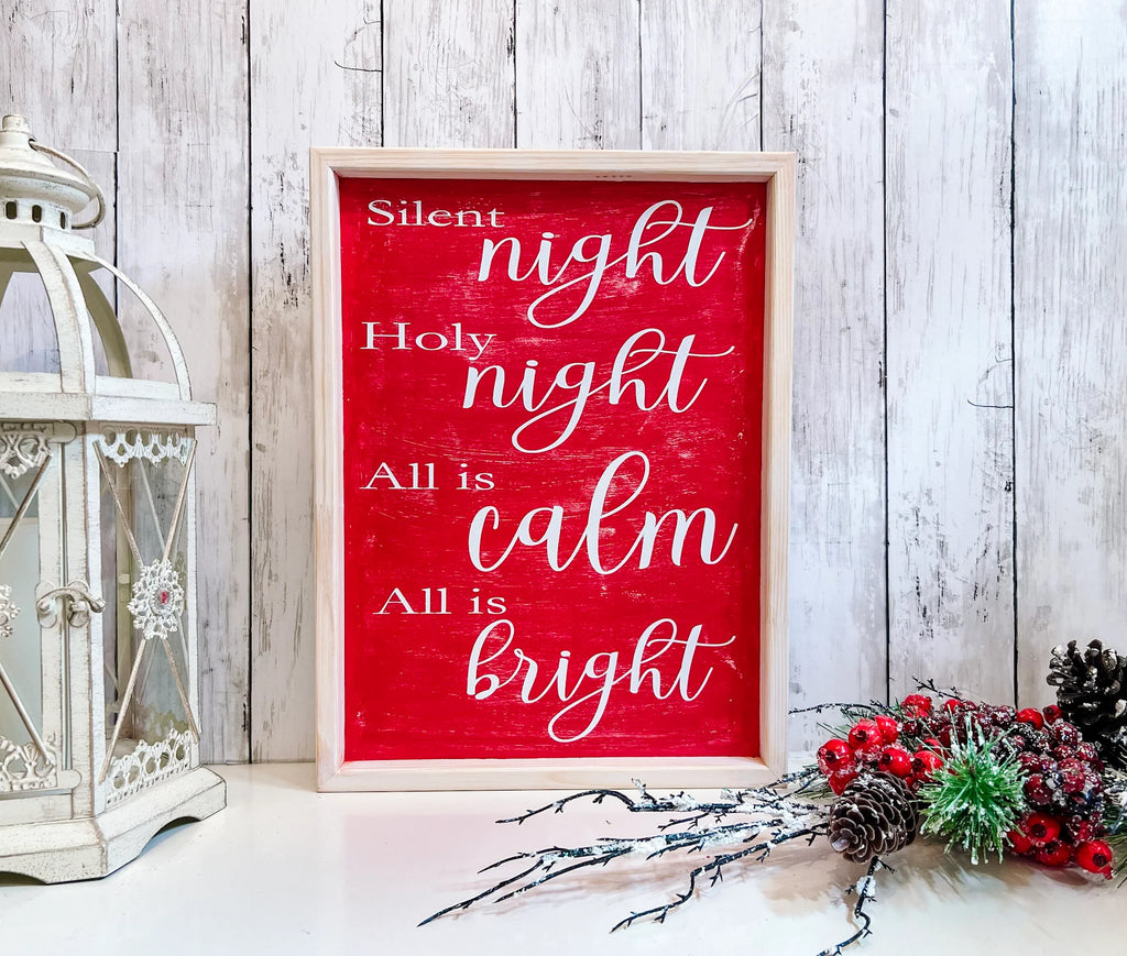 Red Silent Night Holy Night All is Calm All is Bright framed wood sign with whitewashed birch frame leaning against white shiplap wall with red berry pine cone spray angled view