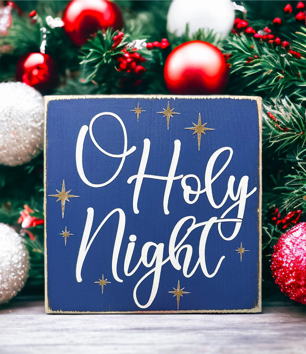 Oh Holy Night navy blue wood block sign with white script and gold stars in front of Christmas tree with warm bokeh lights close up