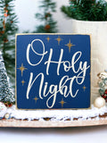 Oh Holy Night navy blue wood block sign with white script and gold stars on white wood surface with pine cones angled view