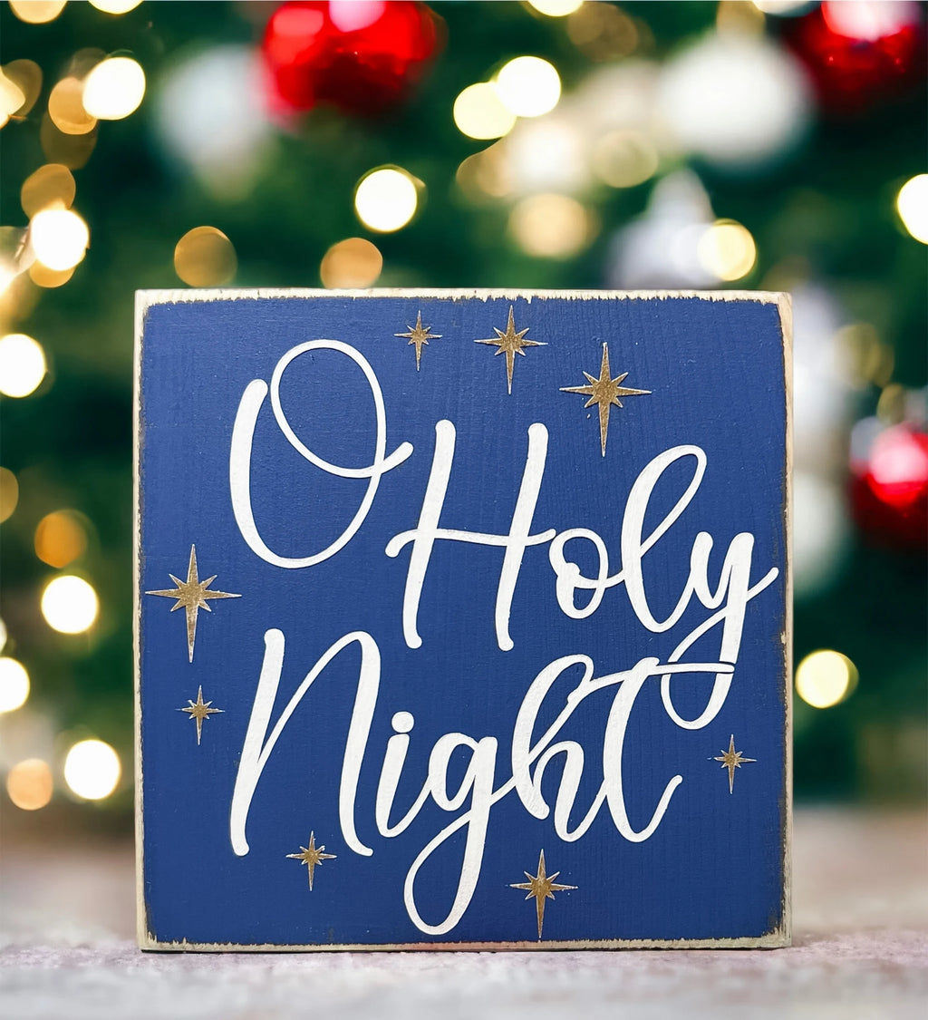 Oh Holy Night navy blue wood block sign with white script and gold stars on white wood surface with pine cones and Christmas greenery