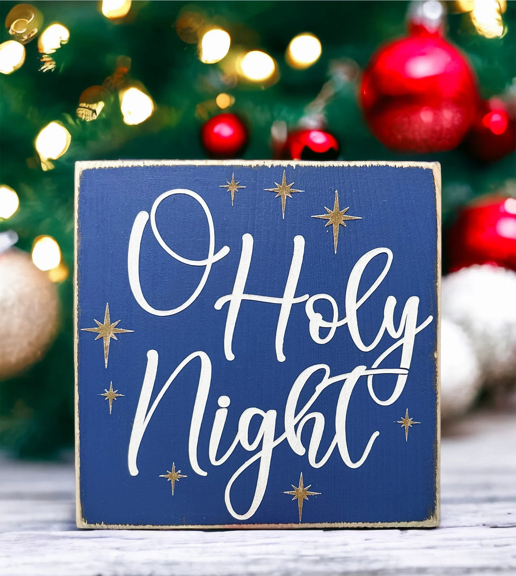 Oh Holy Night navy blue wood block sign with white script and gold stars on white wood surface with pine cones and warm bokeh lights