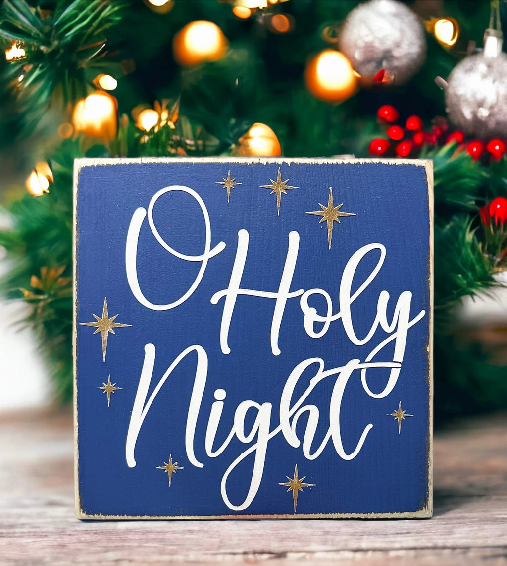 Oh Holy Night navy blue wood block sign with white script and gold stars on snowy wood surface with pine cones and Christmas tree background