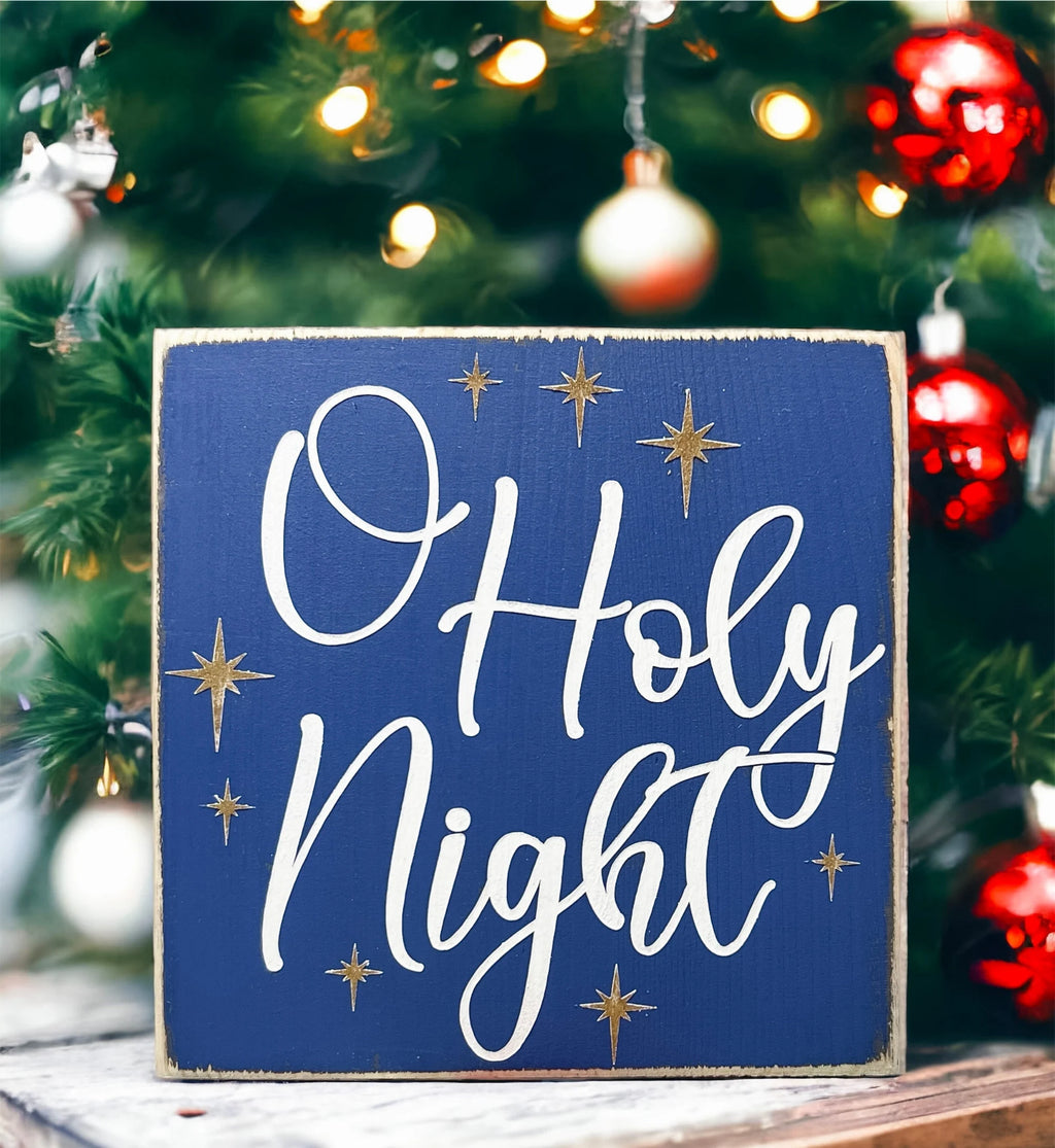 Oh Holy Night navy blue wood block sign with white script and gold stars in front of Christmas tree with warm bokeh lights wide shot