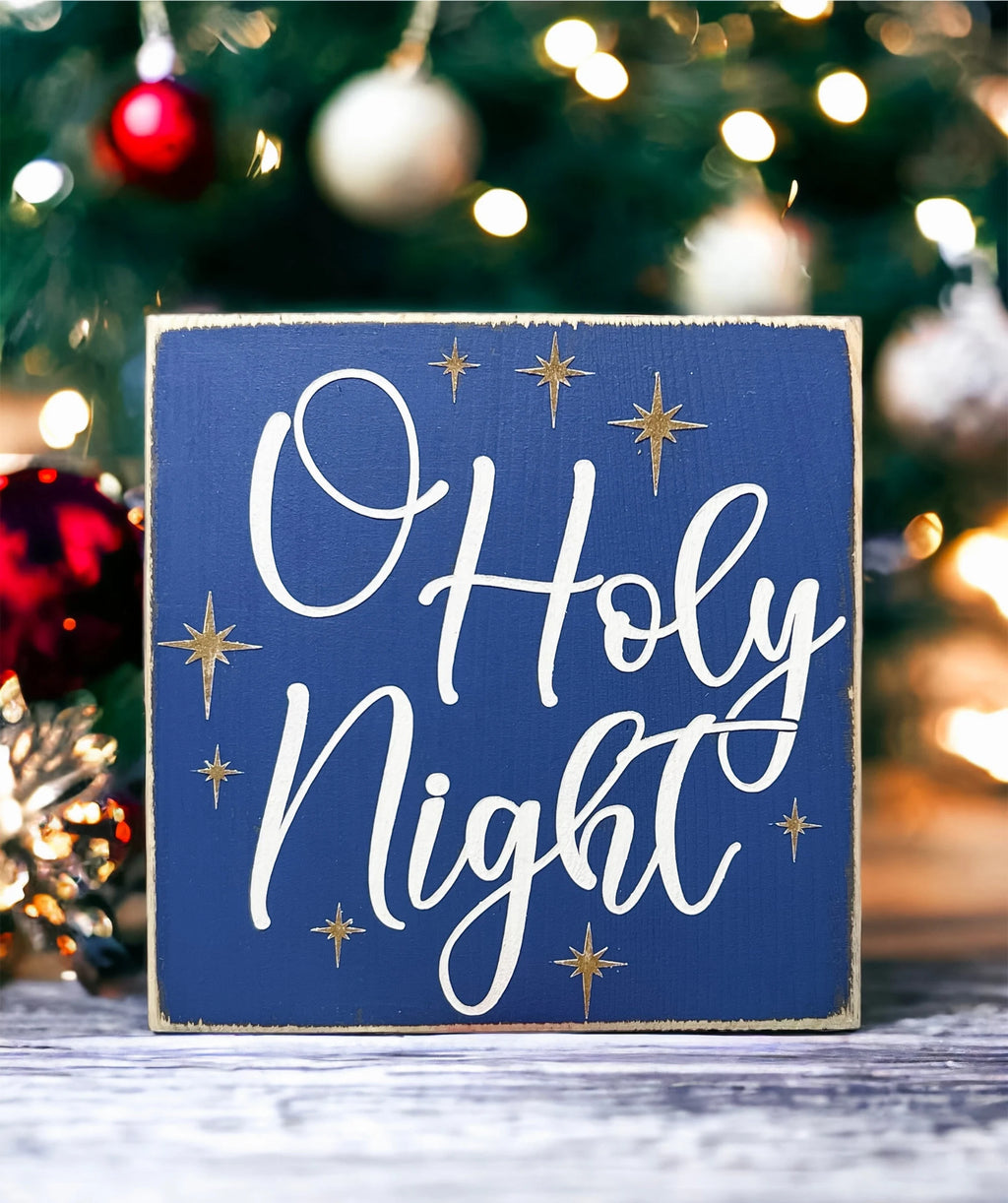 Oh Holy Night navy blue wood block sign with white script and gold stars in front of Christmas tree with red ornaments bokeh background