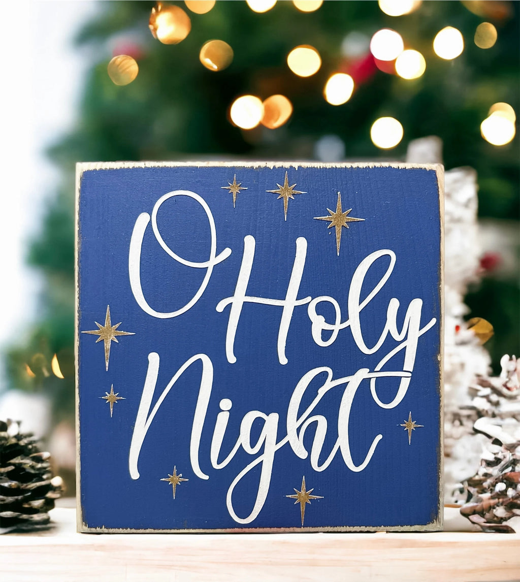Oh Holy Night navy blue wood block sign with white script and gold stars on white wood surface with pine cones product shot