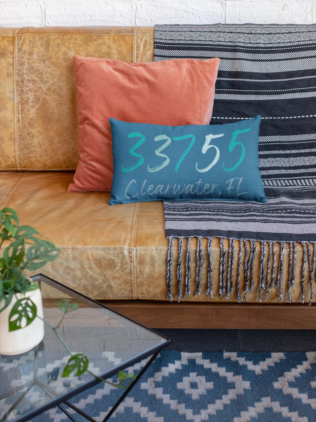 Personalized 33755 Clearwater FL navy lumbar pillow on white wicker chair with cream knit blanket and tropical plant lifestyle shot
