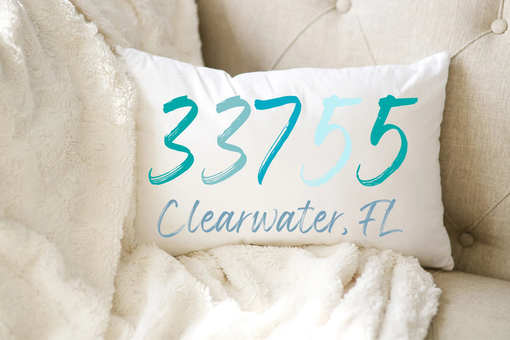 Personalized zip code lumbar pillow reading 33755 Clearwater FL in coastal teal blues on white distressed wood chair in bright window light