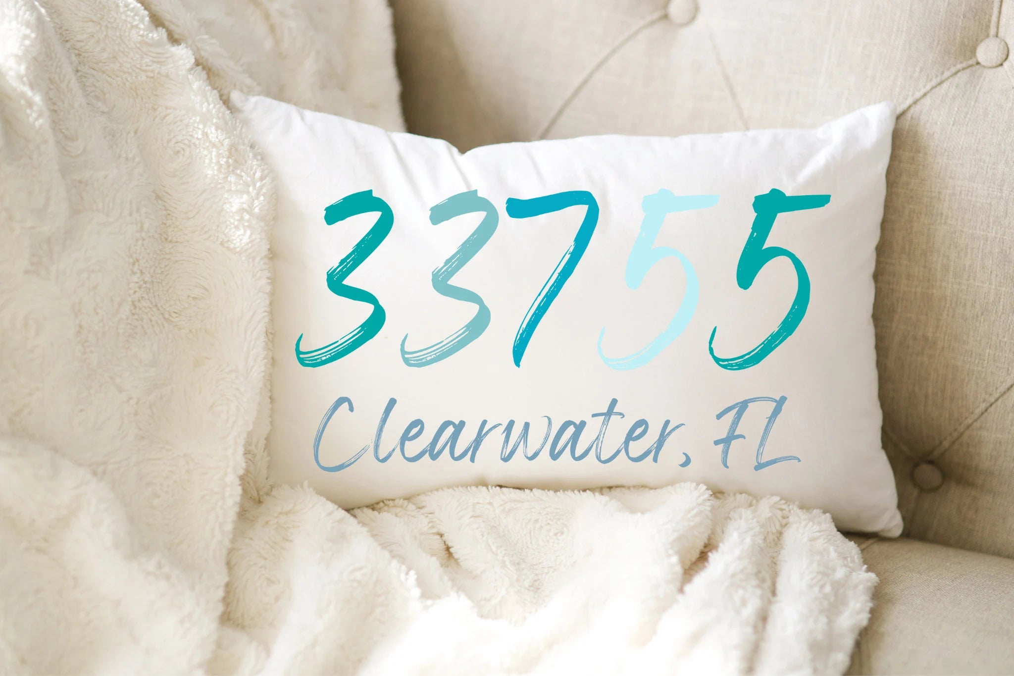 Personalized zip code lumbar pillow reading 33755 Clearwater FL in coastal teal blues on white distressed wood chair in bright window light
