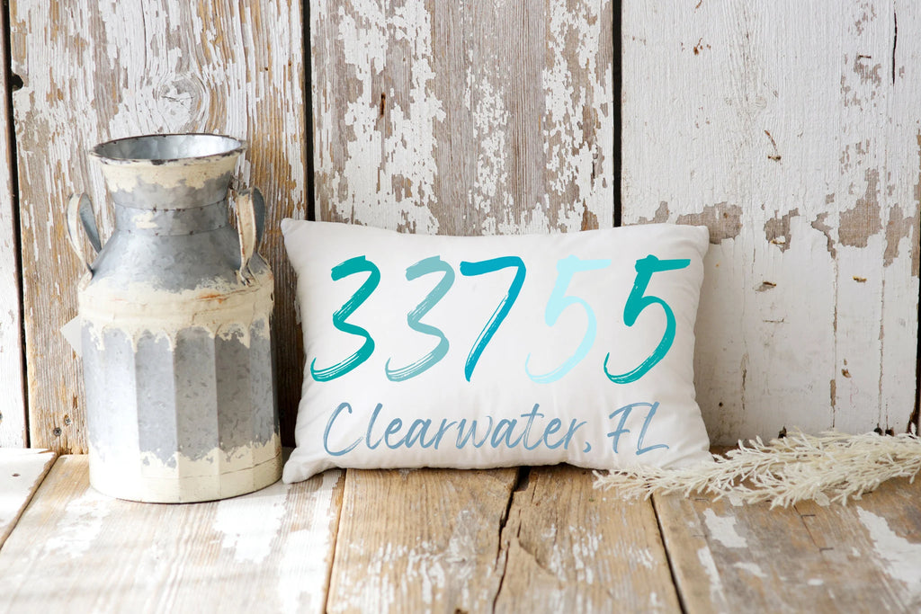 Personalized zip code lumbar pillow reading 33755 Clearwater FL in coastal teal blues on cream tufted sofa with gray knit blanket close up