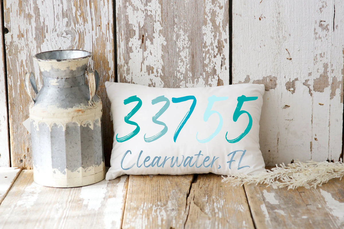 Personalized zip code lumbar pillow reading 33755 Clearwater FL in coastal teal blues on cream tufted sofa with gray knit blanket close up