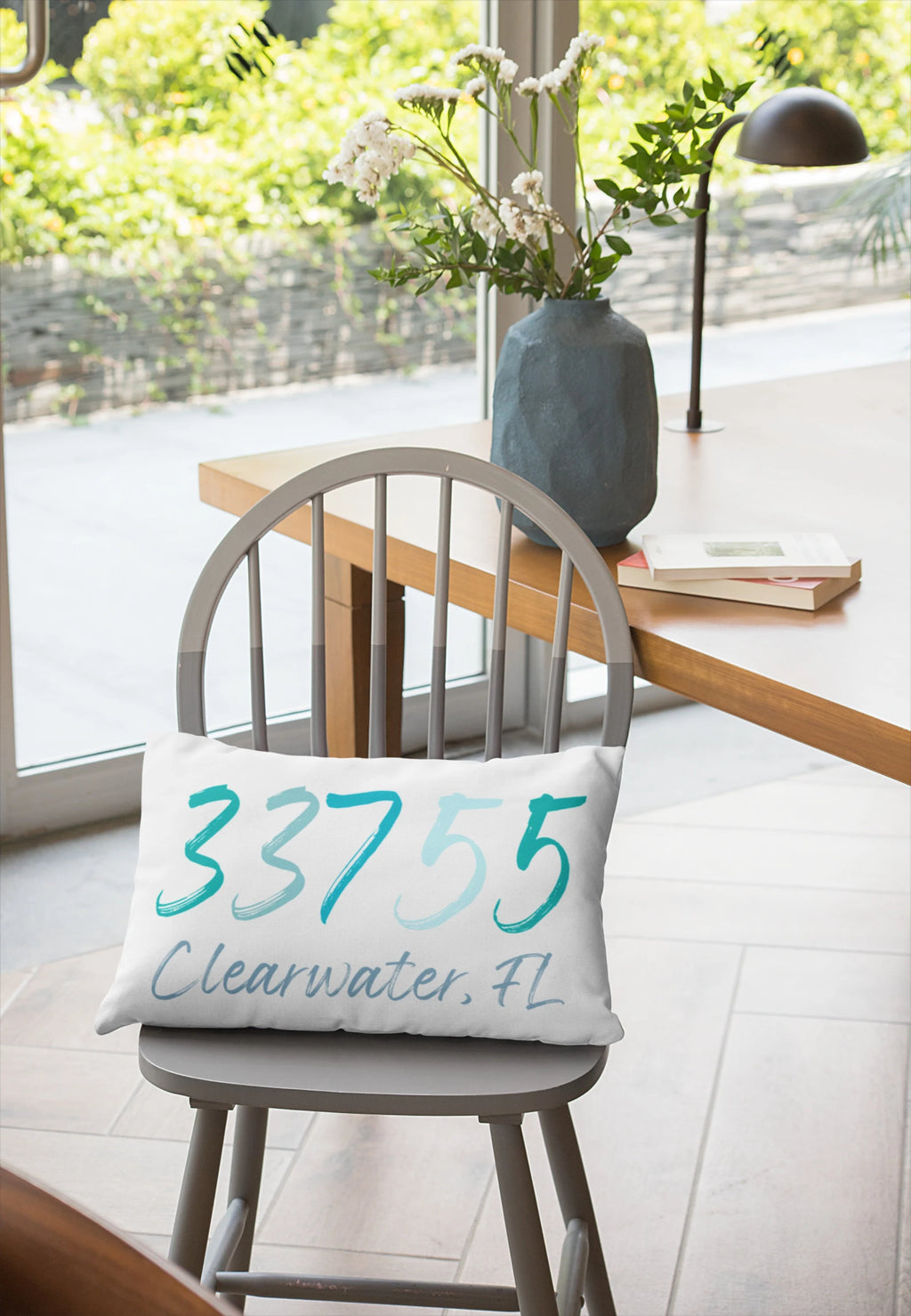 Personalized zip code lumbar pillow reading 33755 Clearwater FL in coastal teal blues leaning against white distressed wood wall on rustic floor