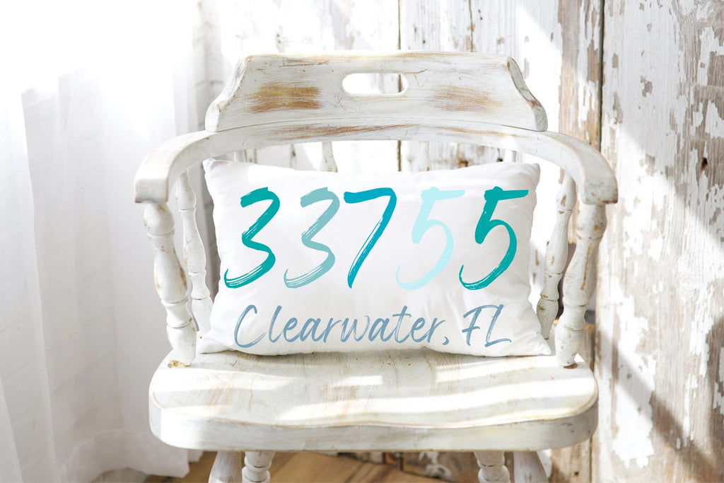Personalized zip code lumbar pillow reading 33755 Clearwater FL in coastal teal blues on rustic wood floor with galvanized milk jug wide shot