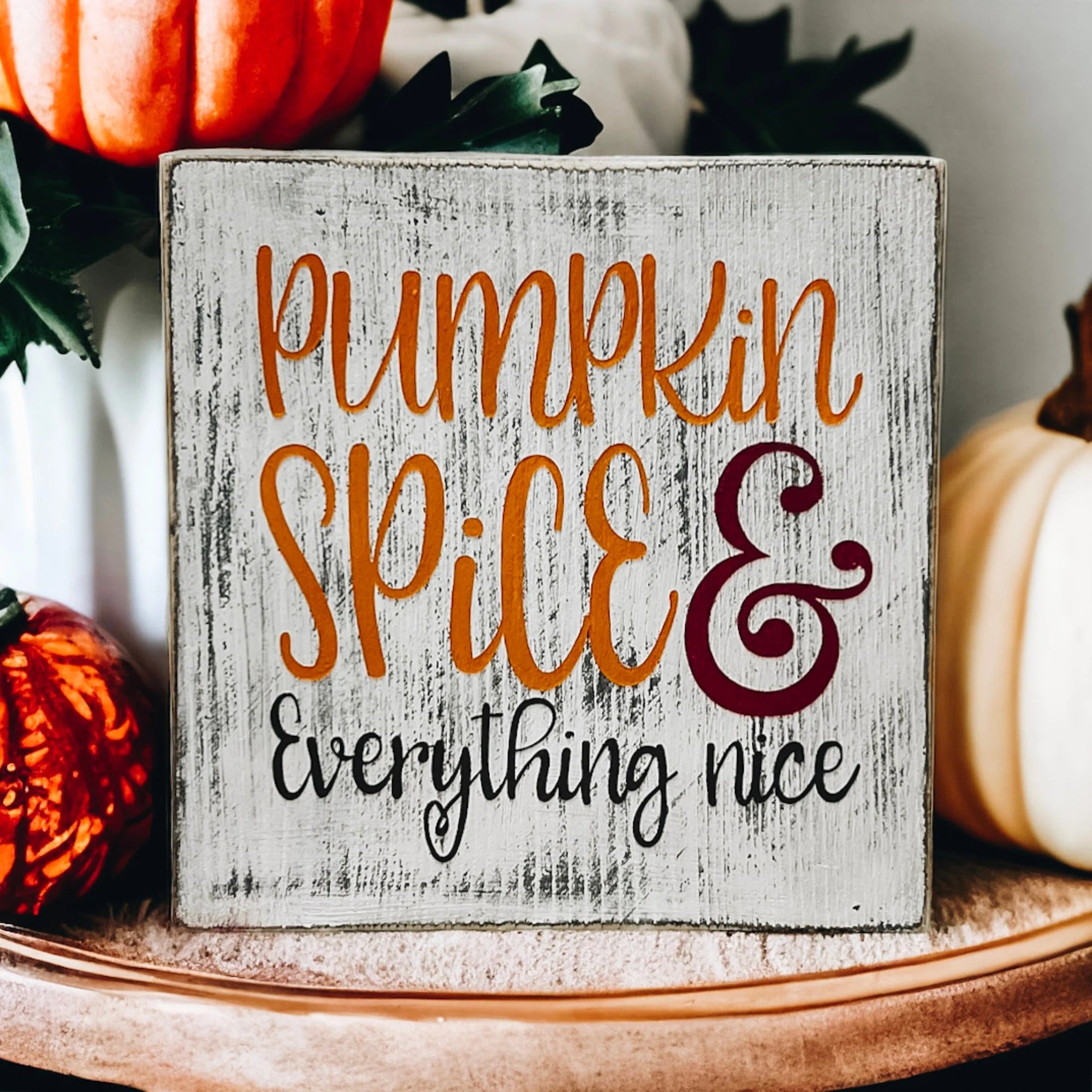 Pumpkin Spice and Everything Nice distressed whitewash wood block sign with amber script orange serif and burgundy ampersand on rustic wood surface with orange pumpkins and greenery angled view