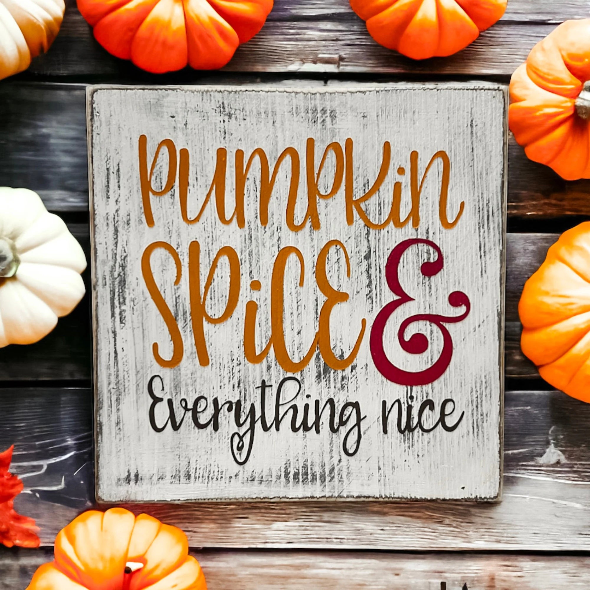 Pumpkin Spice and Everything Nice distressed whitewash wood block sign with amber script orange serif and burgundy ampersand surrounded by orange and white pumpkins on rustic wood surface close up