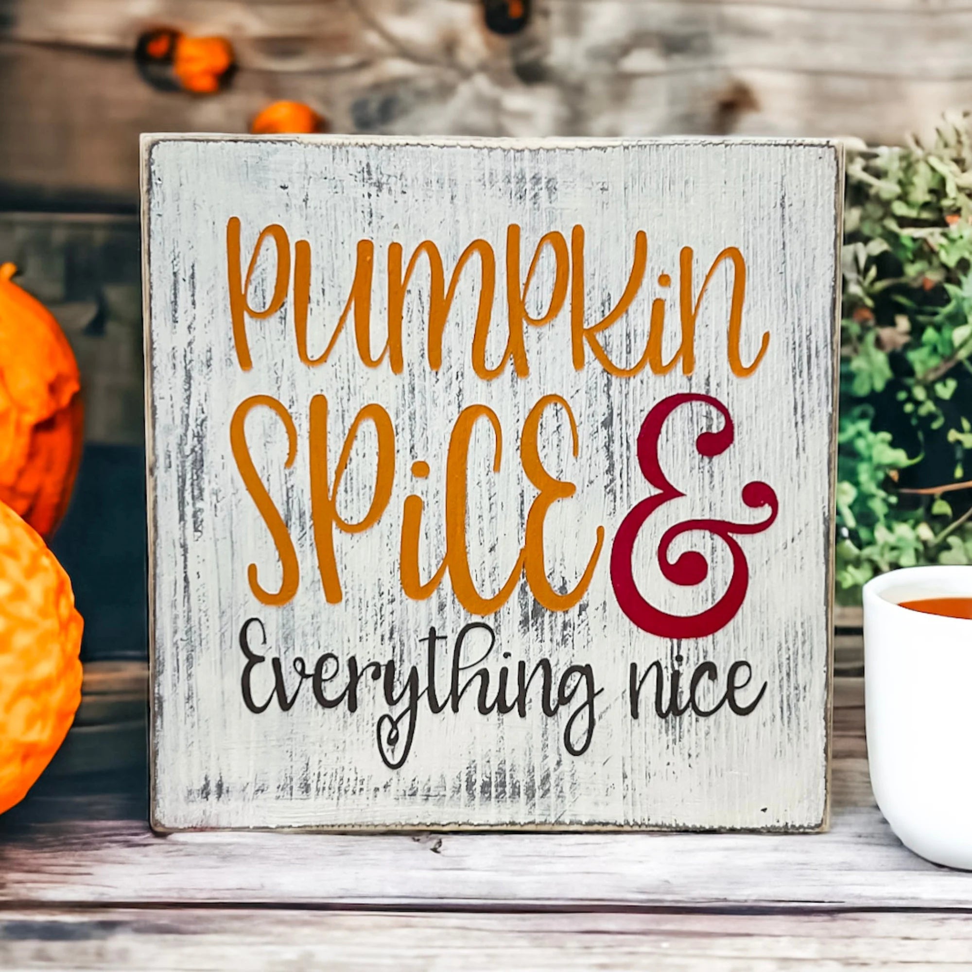 Pumpkin Spice and Everything Nice distressed whitewash wood block sign with amber script orange serif and burgundy ampersand leaning against orange pumpkins on rustic wood surface