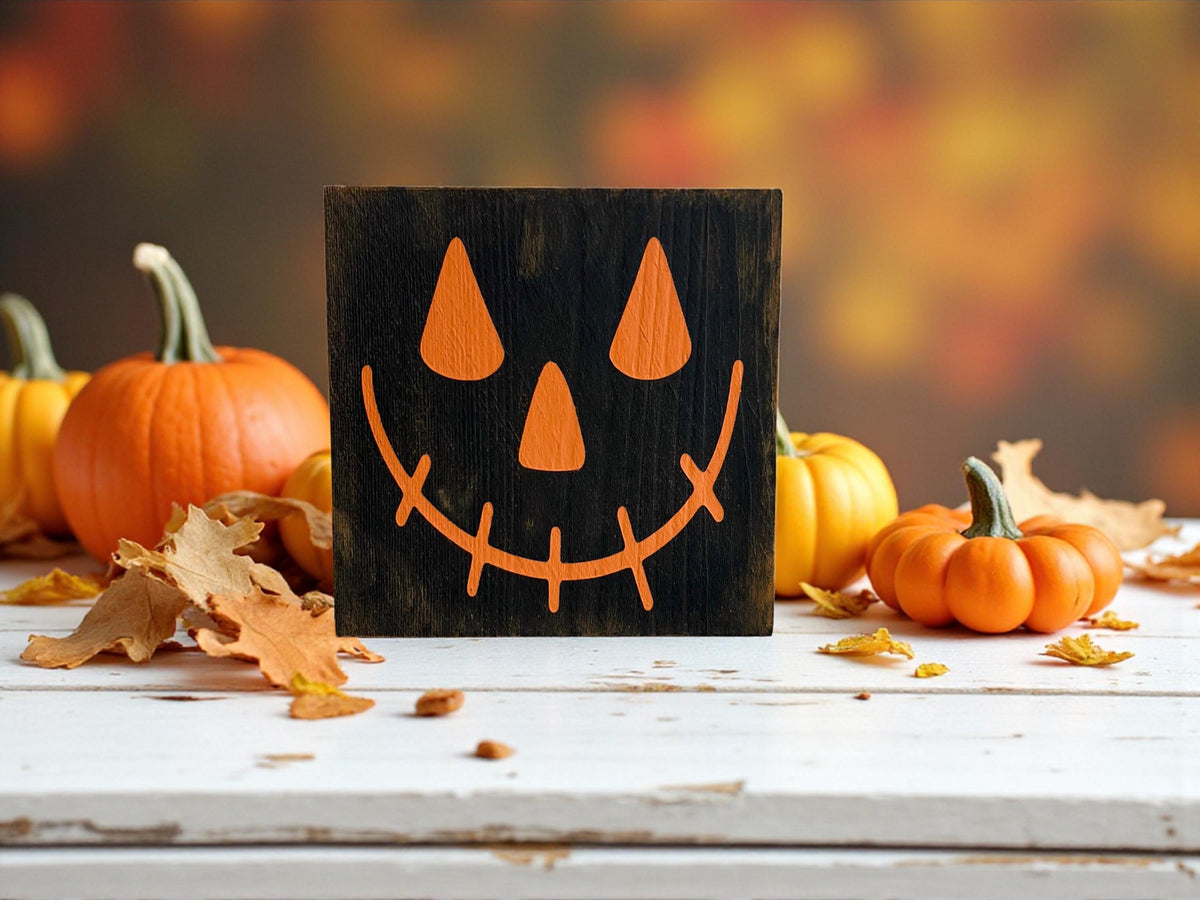 Jack-o-lantern black wood block sign with orange face on rustic wood shelf with orange pumpkins and fall foliage background