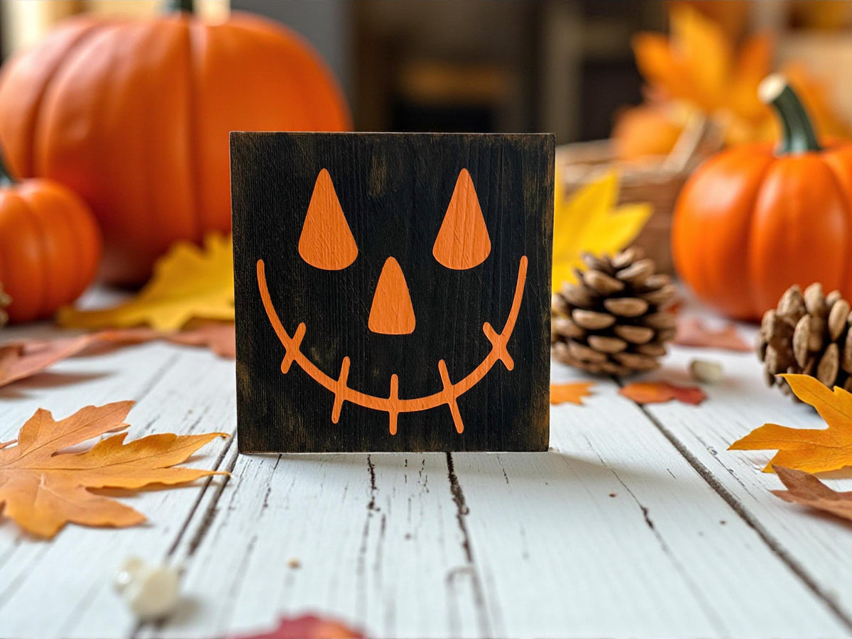 Jack-o-lantern black wood block sign with orange face on white marble counter with orange and white pumpkins and red fall leaves
