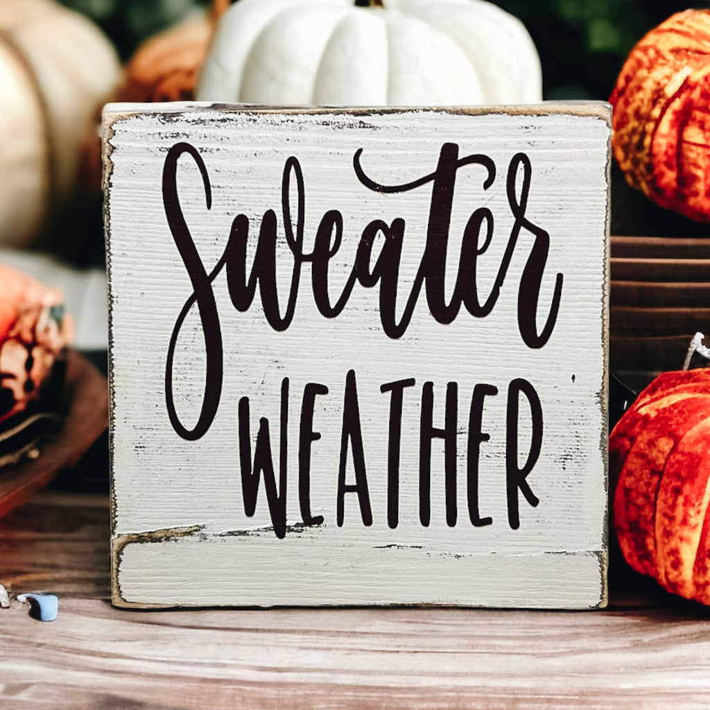Sweater Weather sign on white surface with fall flowers and pumpkin