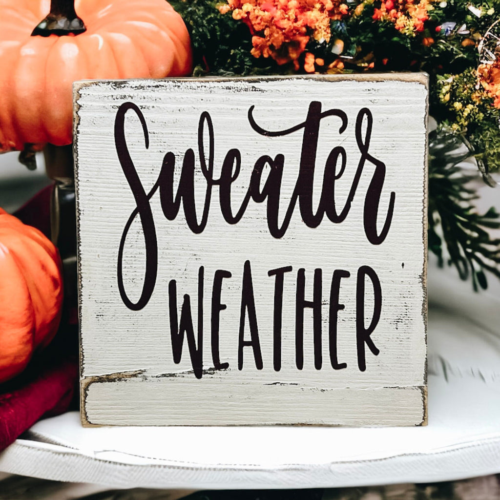 Sweater Weather sign propped on rustic wood with orange and white pumpkins