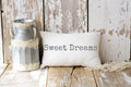 Sweet Dreams white lumbar pillow with gray distressed typewriter lettering on white knit throw blanket in wicker basket lifestyle shot angled