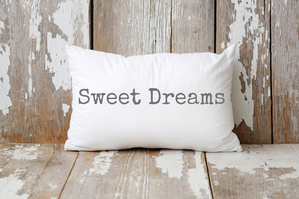 Sweet Dreams white lumbar pillow with gray distressed typewriter lettering leaning against peeling white barn wood wall on rustic wood floor hero shot