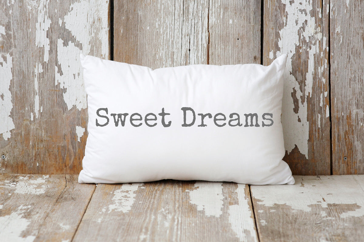 Sweet Dreams white lumbar pillow with gray distressed typewriter lettering leaning against peeling white barn wood wall on rustic wood floor hero shot