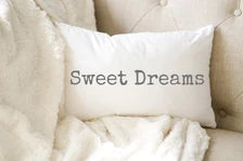 Sweet Dreams white lumbar pillow with gray distressed typewriter lettering leaning against peeling white barn wood wall on rustic wood floor with vintage galvanized milk jug angled view