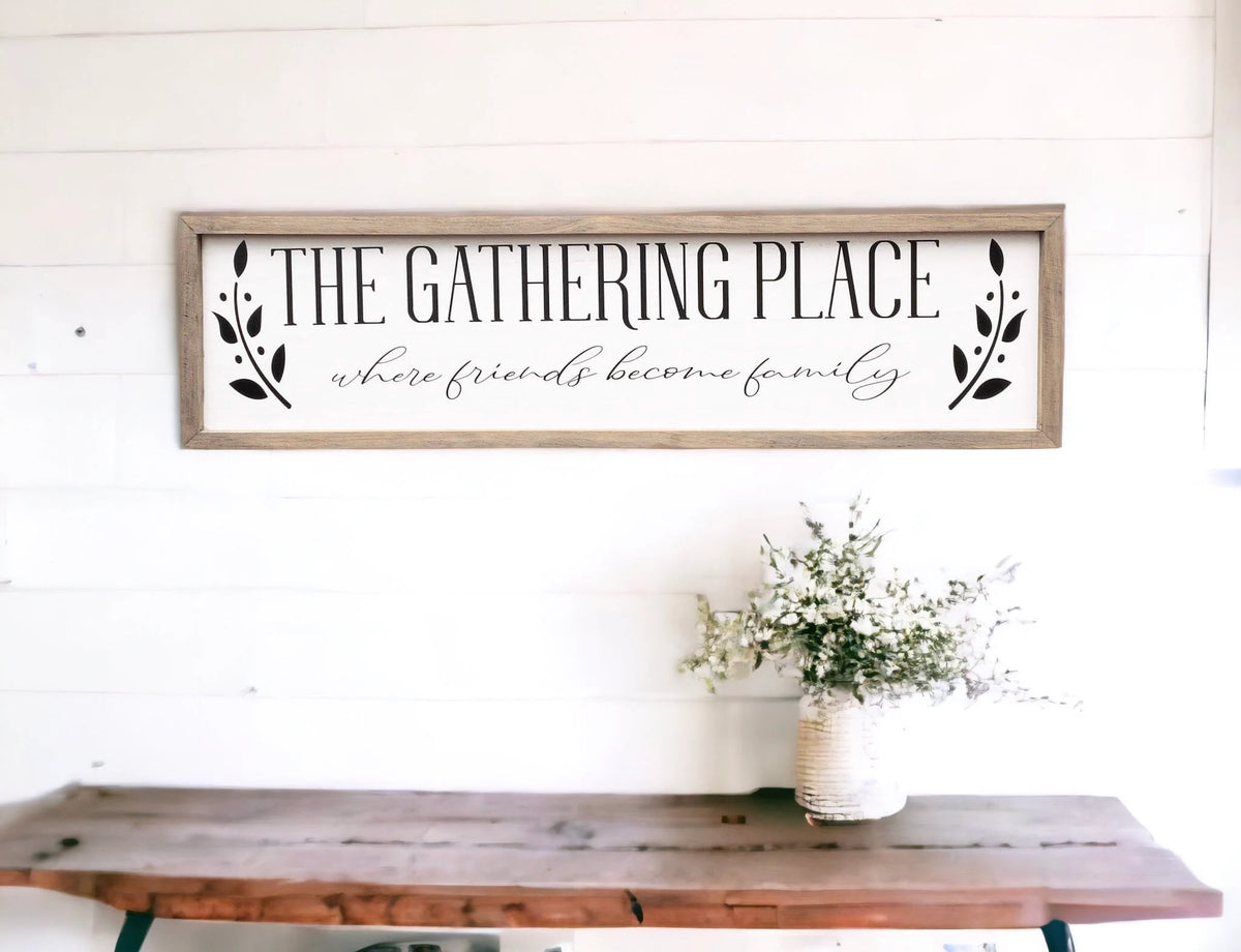 The Gathering Place where friends become family framed farmhouse sign on white shiplap wall above rustic wood console table close up