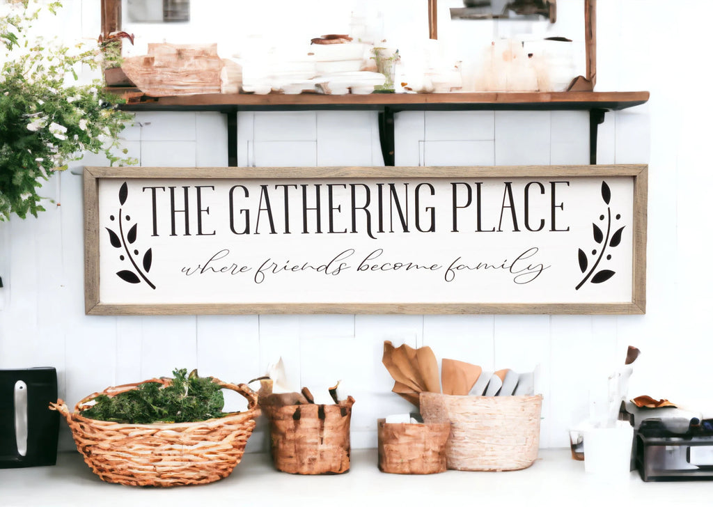 The Gathering Place where friends become family framed farmhouse sign on white shiplap wall above wood bench with wicker basket and greenery wide shot
