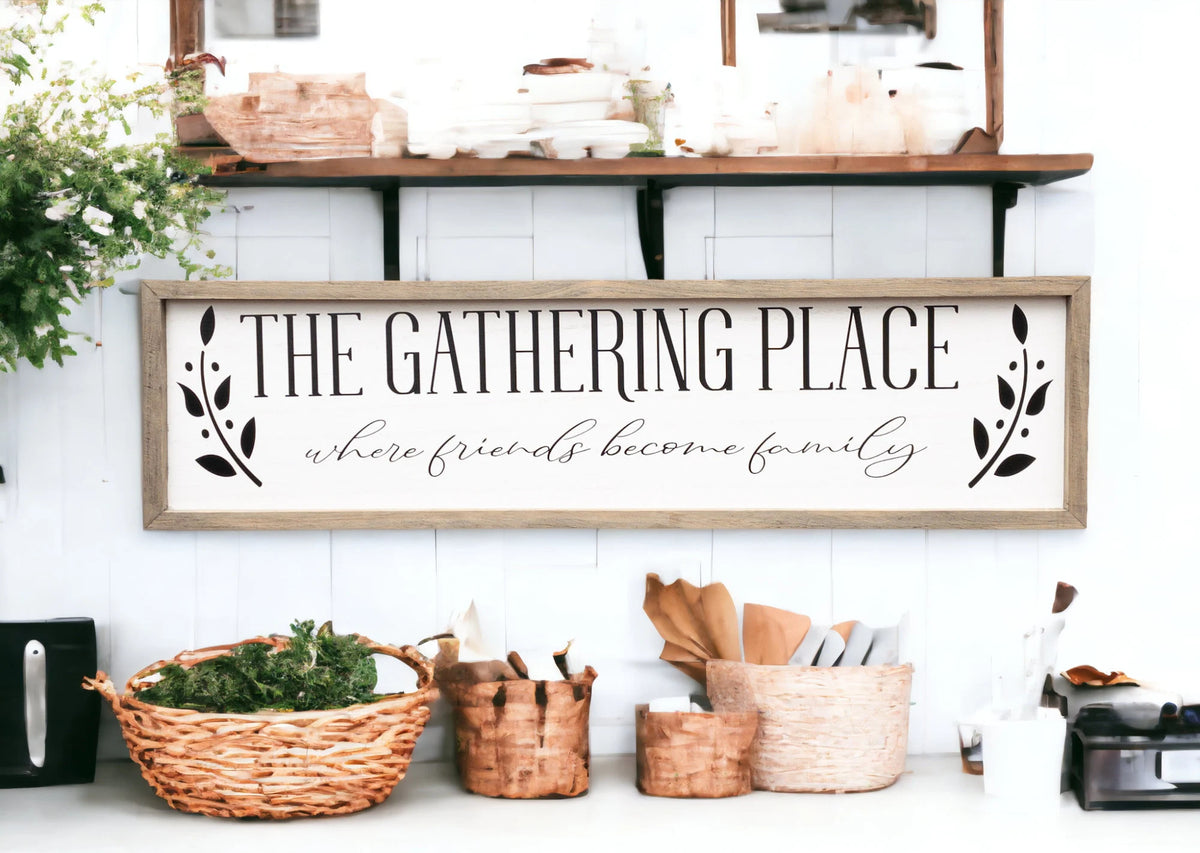 The Gathering Place where friends become family framed farmhouse sign on white shiplap wall above wood bench with wicker basket and greenery wide shot