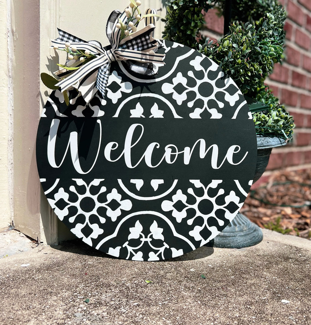Black and white tile pattern Welcome round door hanger leaning on white door frame with boxwood topiary