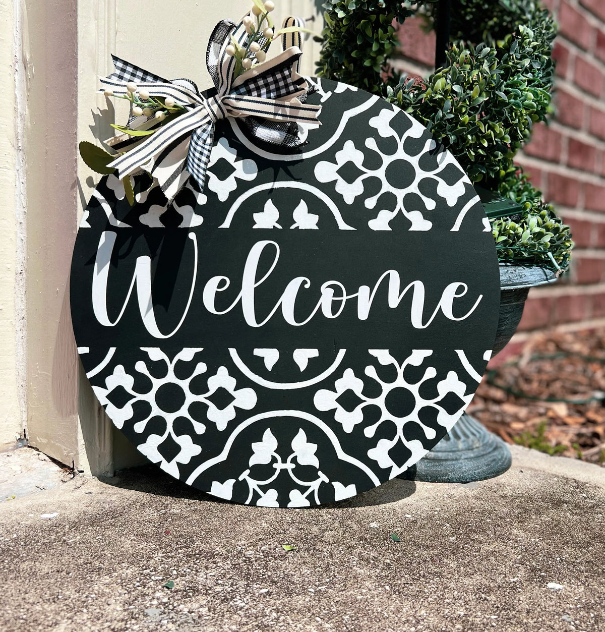 Black and white tile pattern Welcome round door hanger leaning on white door frame with boxwood topiary