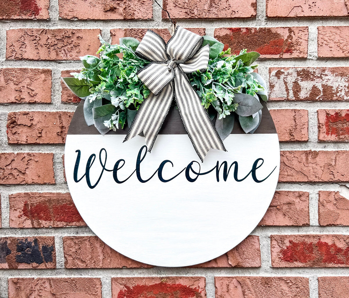 Farmhouse Welcome round door hanger with ticking stripe bow and eucalyptus greenery on brick wall full view