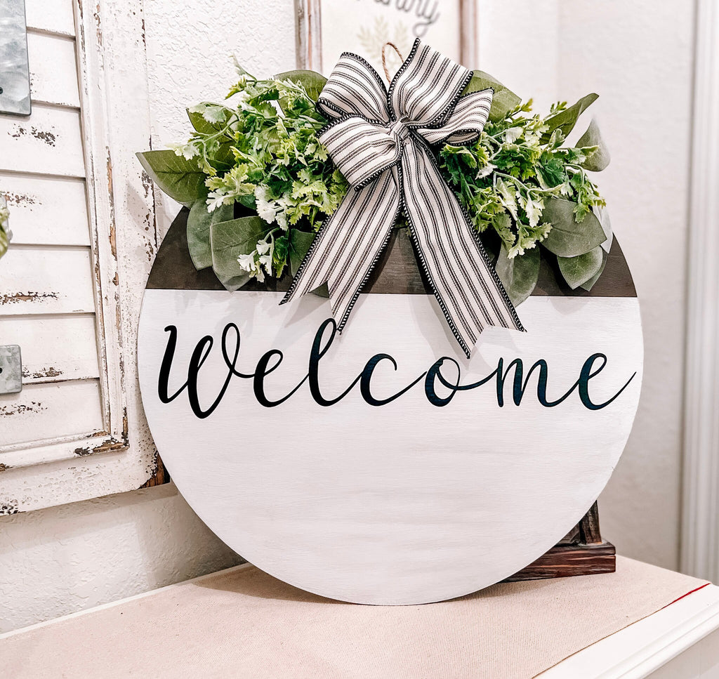 Farmhouse Welcome round door hanger with ticking stripe bow and eucalyptus greenery on white shutter shelf close up