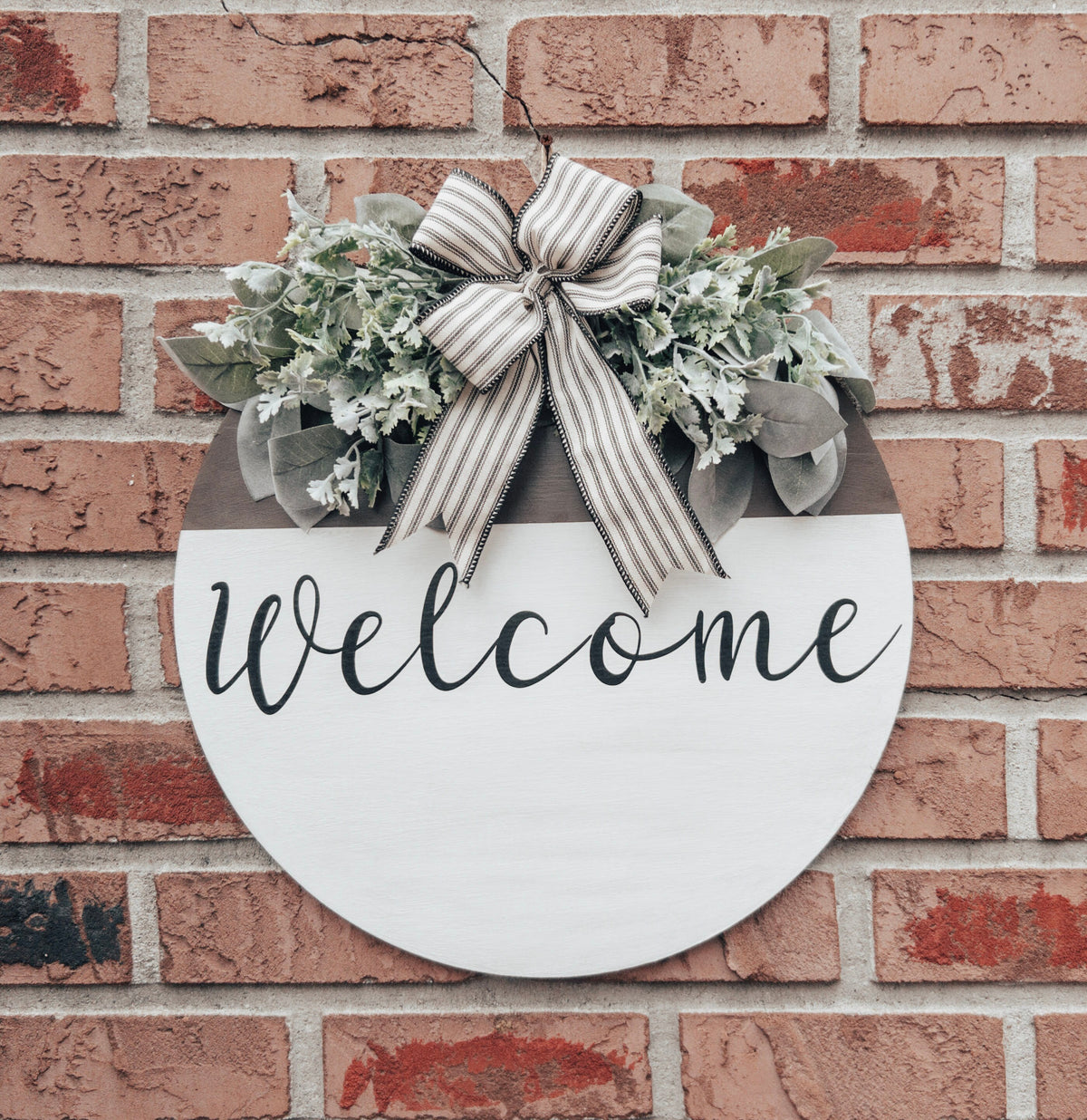 Farmhouse Welcome round door hanger with ticking stripe bow and eucalyptus greenery on brick wall angled view