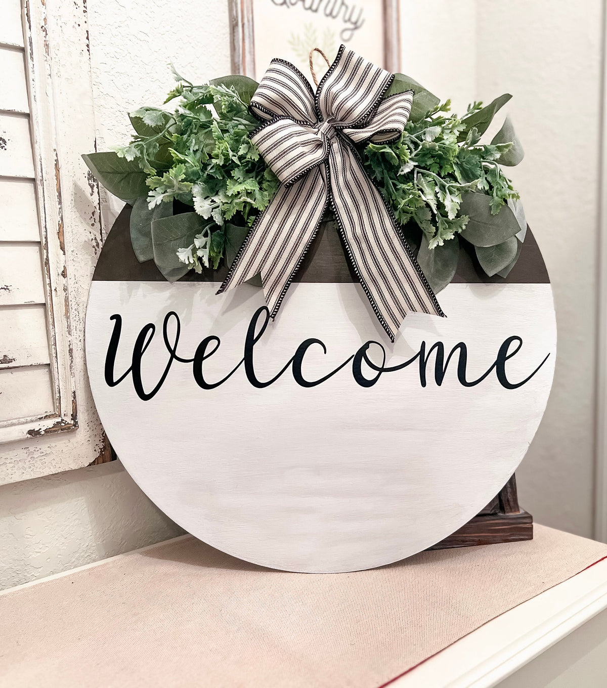 Farmhouse Welcome round door hanger with ticking stripe bow and eucalyptus greenery on white shutter shelf