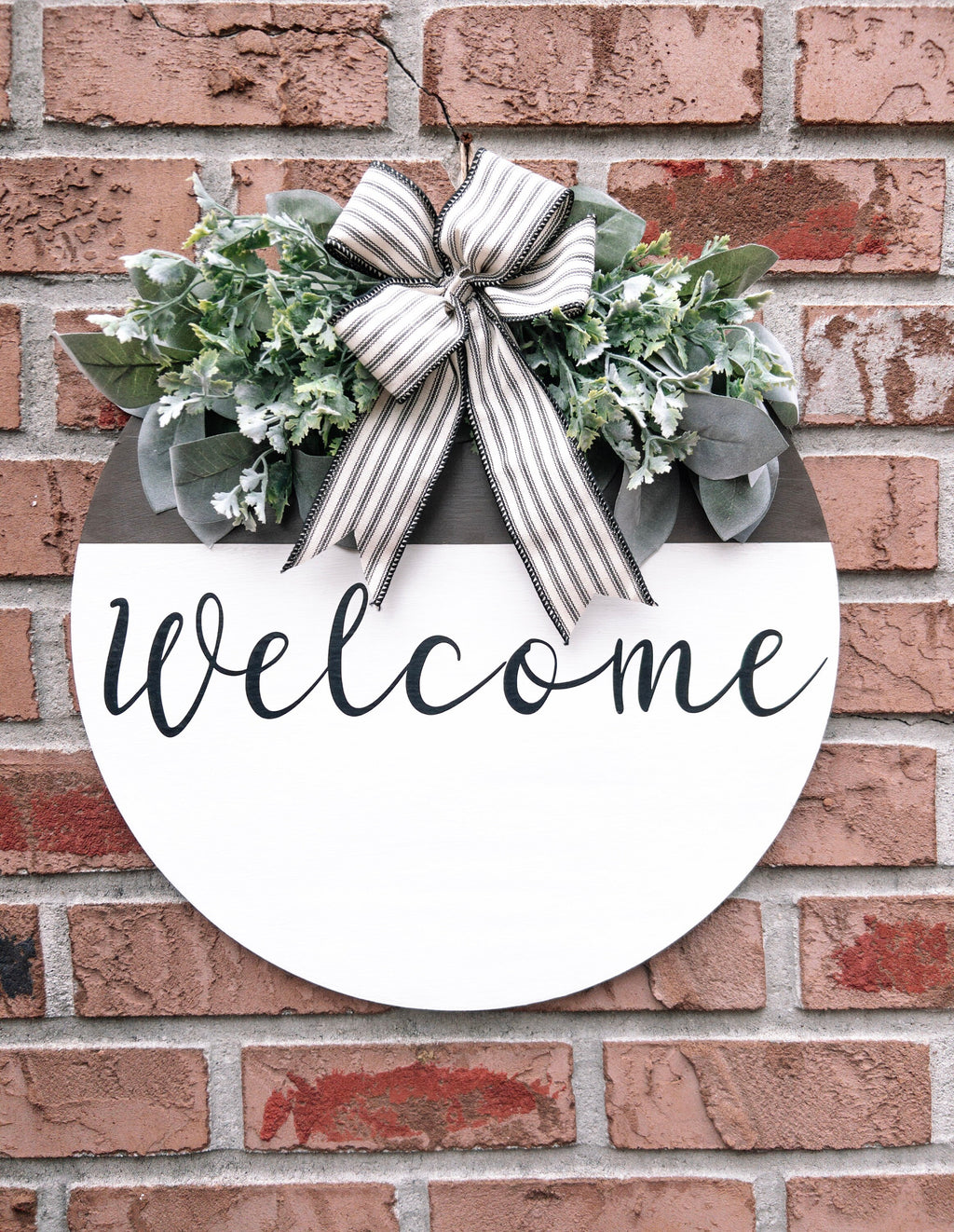 Farmhouse Welcome round door hanger with ticking stripe bow and eucalyptus greenery on brick wall close up