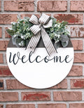 Farmhouse Welcome round door hanger with ticking stripe bow and eucalyptus greenery on brick wall close up