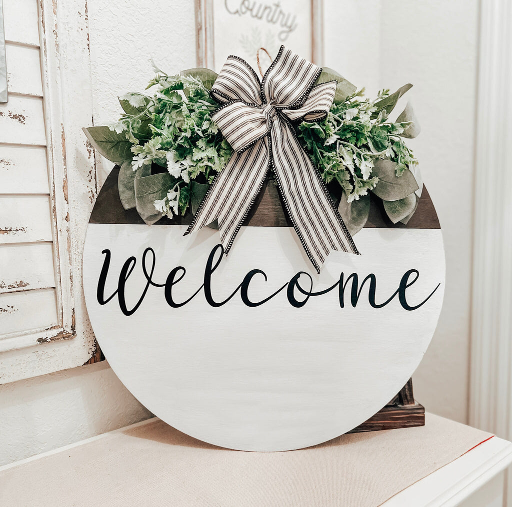 Farmhouse Welcome round door hanger with ticking stripe bow and eucalyptus greenery on white shutter shelf full view