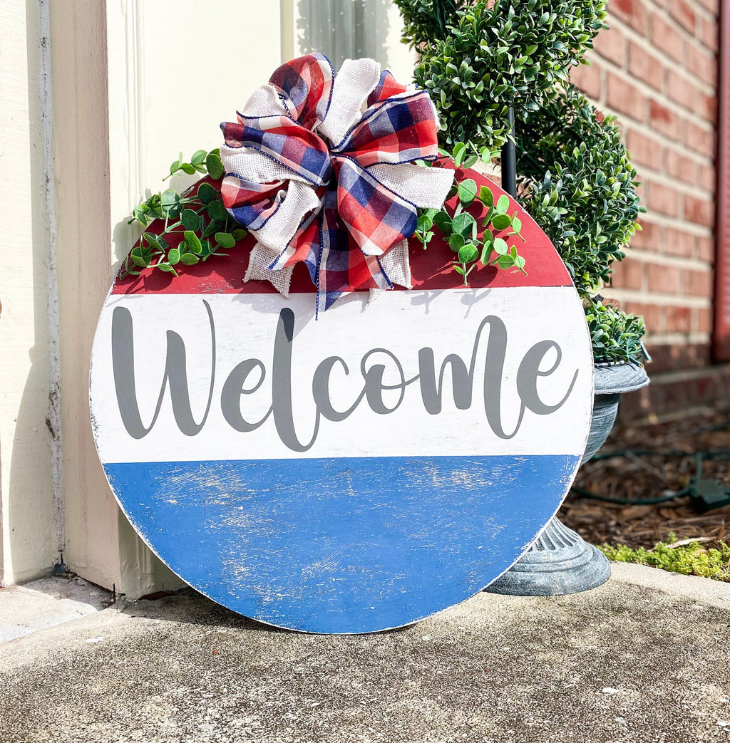 Red white and blue Welcome round pine door hanger with plaid burlap bow and eucalyptus greenery leaning against white door frame with boxwood topiary on brick porch close up bow detail