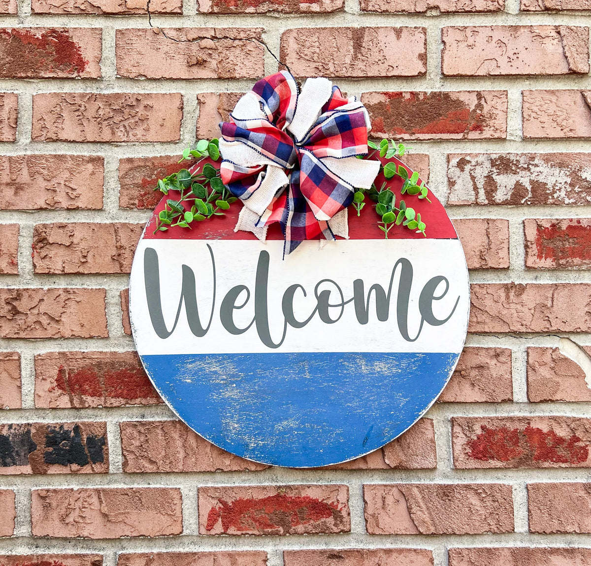 Red white and blue Welcome round pine door hanger with plaid burlap bow and eucalyptus greenery leaning against white door frame with boxwood topiary on brick porch low angle