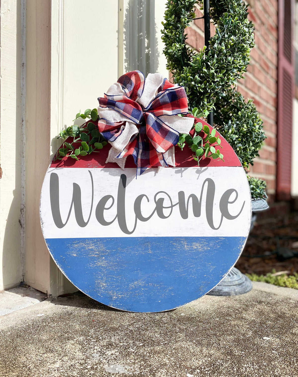 Red white and blue Welcome round pine door hanger with plaid burlap bow and eucalyptus greenery leaning against white door frame with boxwood topiary on brick porch side angle