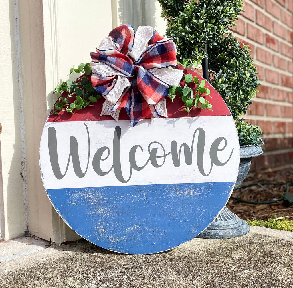 Red white and blue Welcome round pine door hanger with plaid burlap bow and eucalyptus greenery hung on red brick wall front view