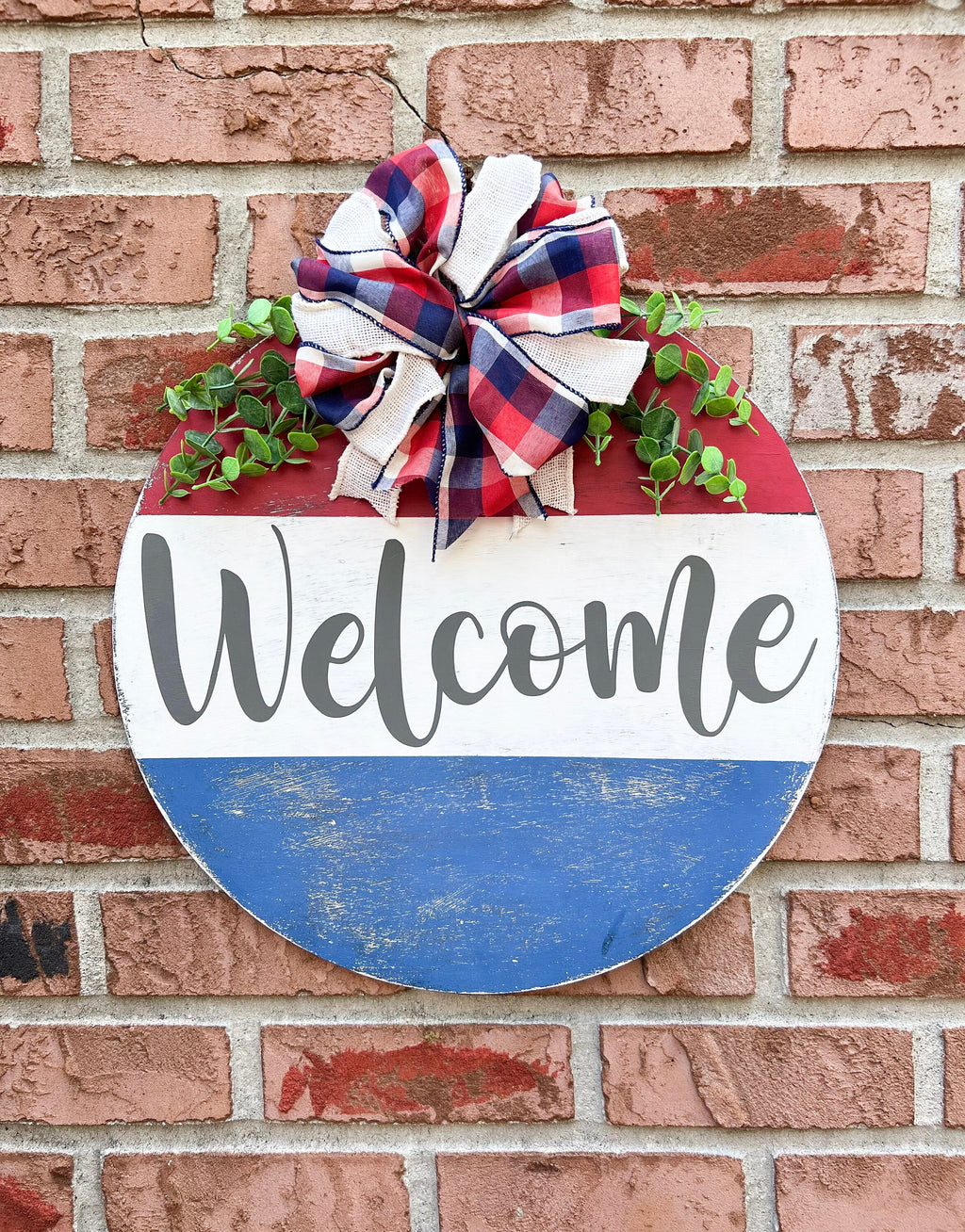Red white and blue Welcome round pine door hanger with plaid burlap bow and eucalyptus greenery leaning against white door frame with boxwood topiary on brick porch angled view