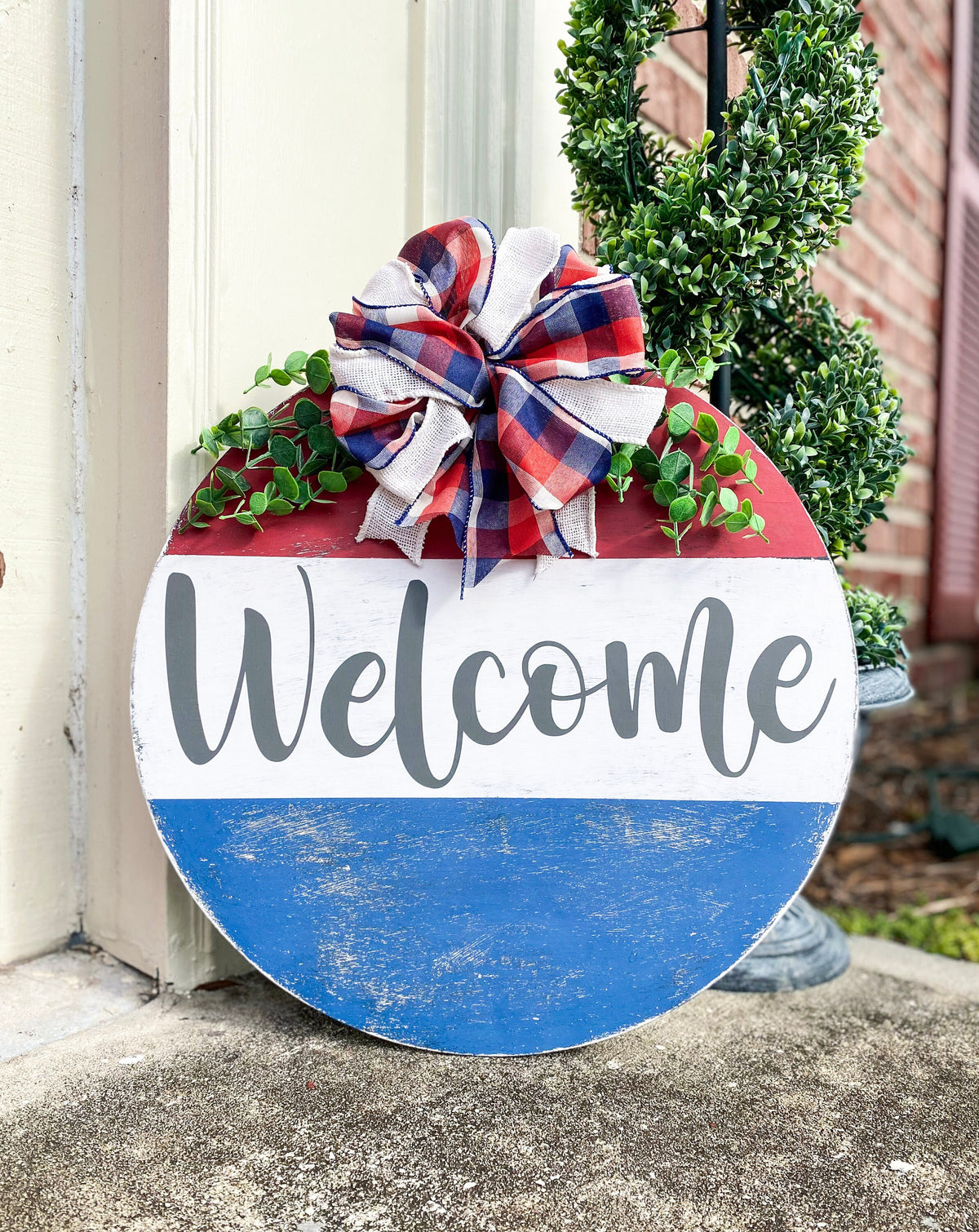 Red white and blue Welcome round pine door hanger with plaid burlap bow and eucalyptus greenery leaning against white door frame with boxwood topiary on brick porch wide shot